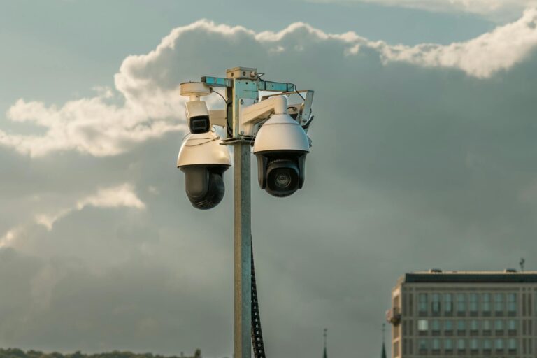Two modern surveillance cameras mounted on a pole in an urban setting under a cloudy sky.